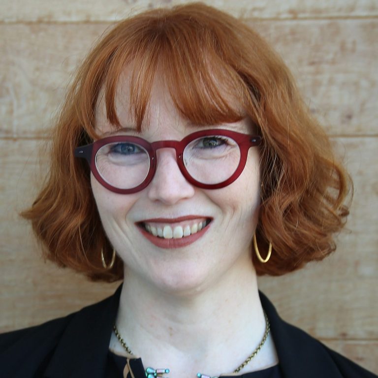 Dr. Dipl.-Psych. Isabella Paul-Jordanov Frau mit rotem, lockigem Haar, Brille und einem freundlichen Lächeln vor einem Holzhintergrund.